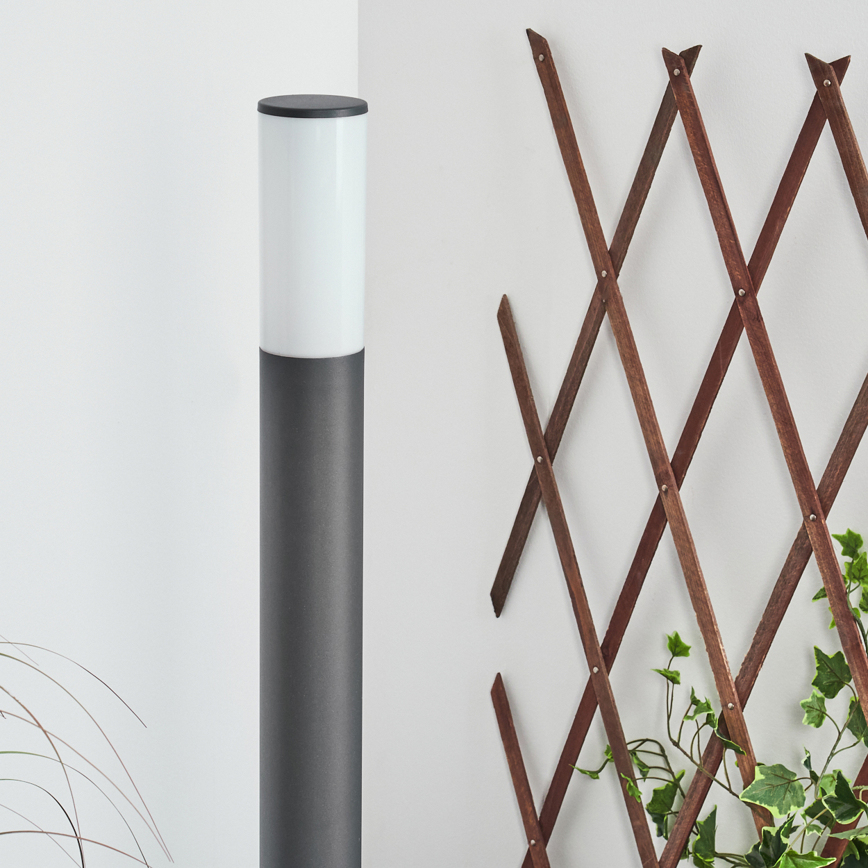 Eine hohe Außenlampe mit weißem Oberteil und grauem Körper, neben einem hölzernen Spalier mit Grünpflanzen.