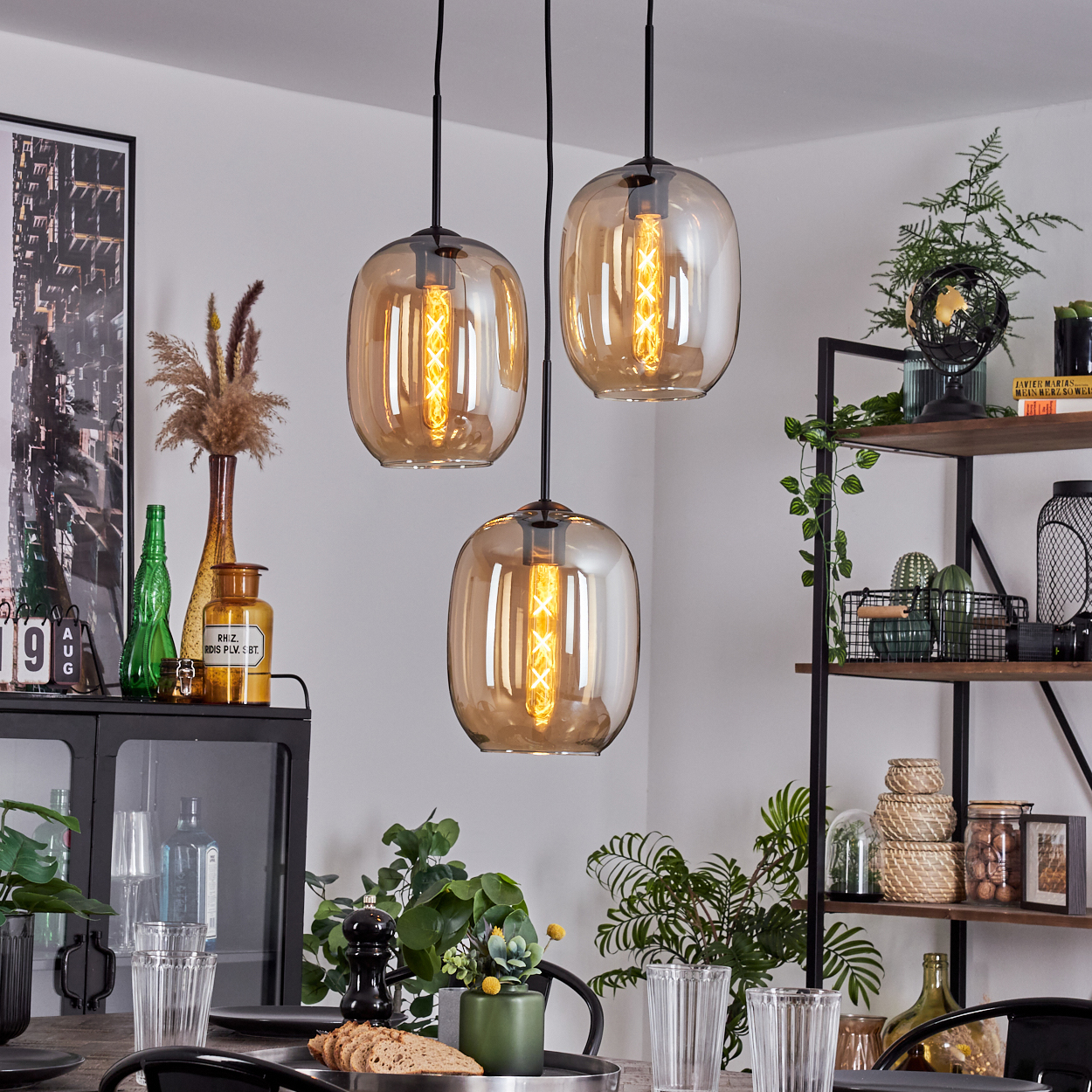 Drei Pendelleuchten mit bernsteinfarbenen Glasschirmen, einer schwarzen Deckenplatte und einem Esszimmer-Setting.