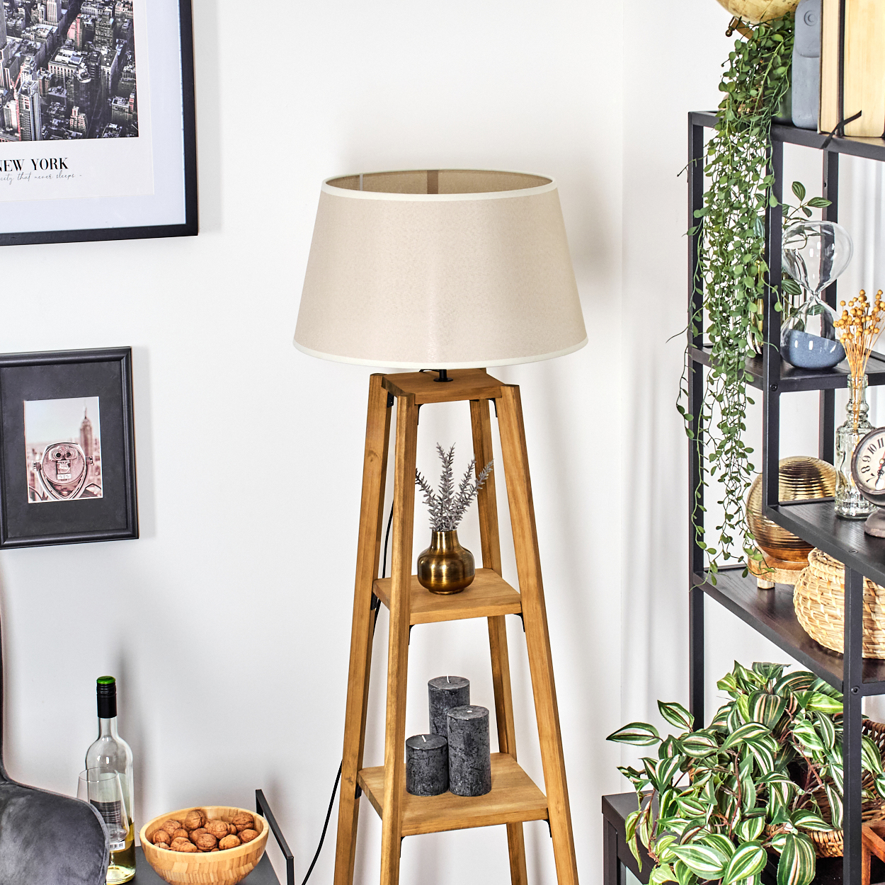 Stehlampe aus Holz mit beigem Lampenschirm. Die Lampe hat Regale mit Dekoration. Hintergrund: Wand, Bilder und Pflanzen.