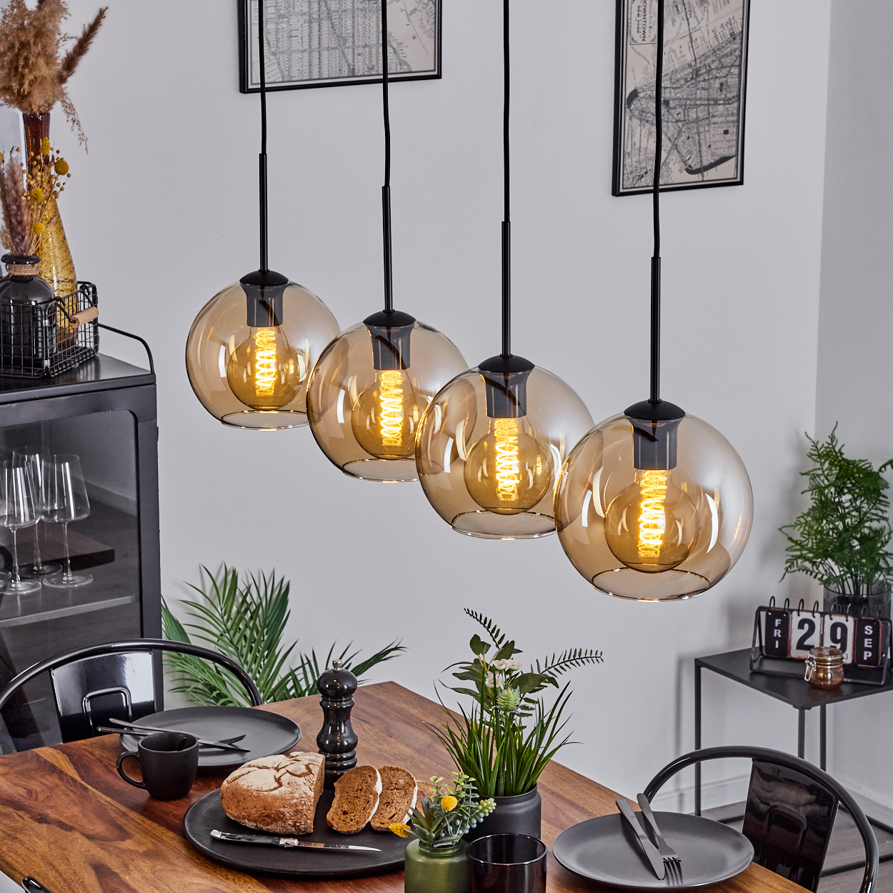 Esszimmer mit Holztisch und vier Pendelleuchten mit bernsteinfarbenen Glühbirnen. Die Einrichtung umfasst Pflanzen und Bilder.