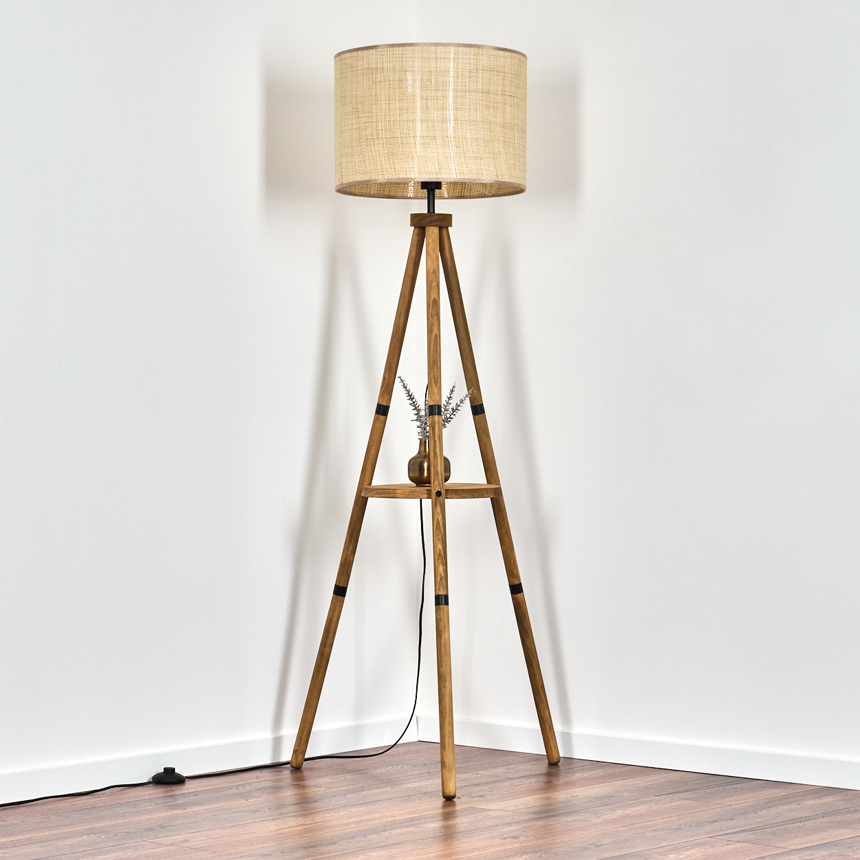Dreibein-Stehleuchte aus Holz in einer Ecke. Beiger Schirm, kleine Vase mit Blumen. Weiße Wände und Holzboden.