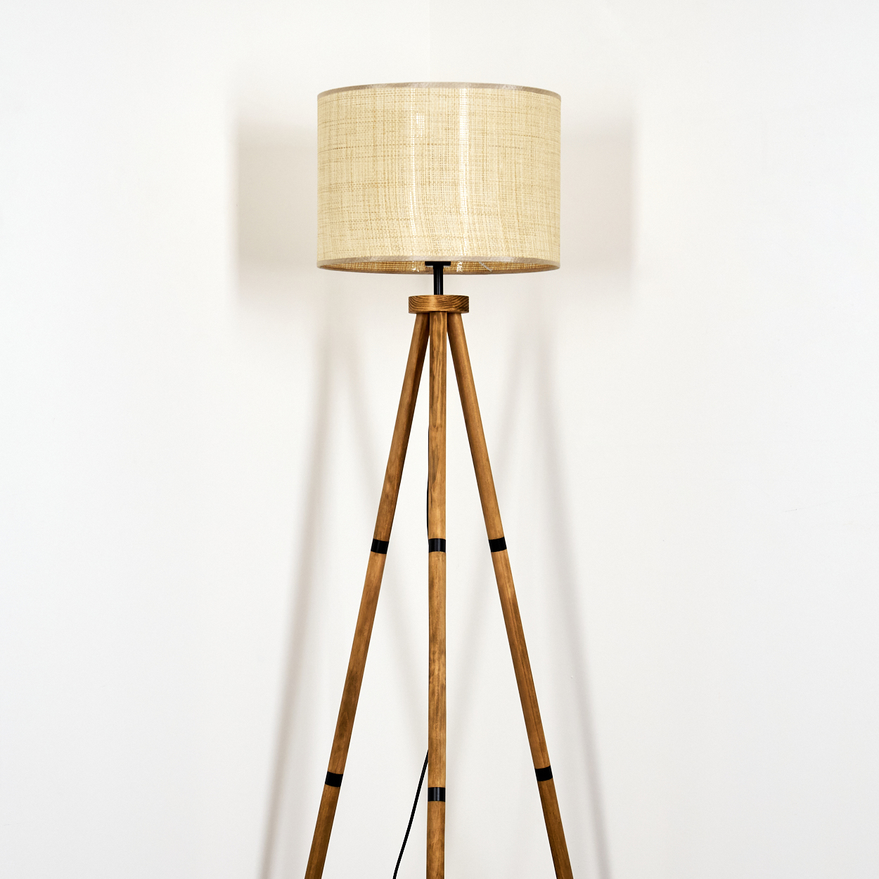 Eine Stehlampe mit beigem Lampenschirm und Holz-Stativbeinen vor einer weißen Wand.