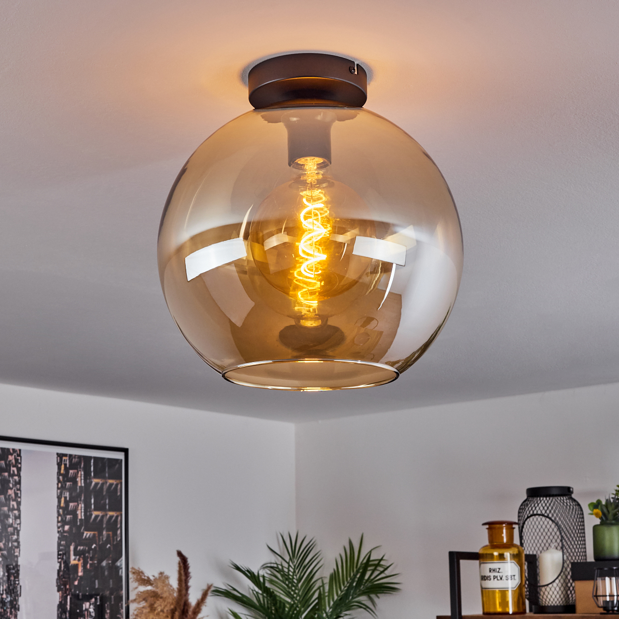 Eine Deckenleuchte mit einem kugelförmigen, bernsteinfarbenen Glasschirm und einer leuchtenden Glühbirne, an einer weißen Decke montiert.
