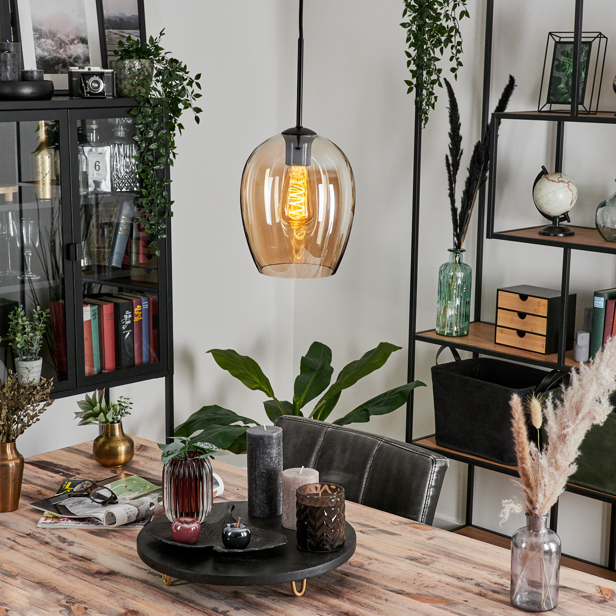Eine einzelne bernsteinfarbene Glas-Pendelleuchte mit einer Edison-Glühbirne. Tisch mit Deko und grünen Pflanzen im Hintergrund.