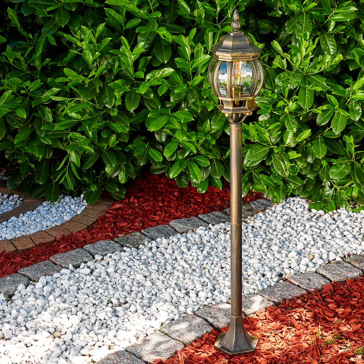 Eine bronzefarbene Straßenlaterne in einem Garten mit grünem Busch, roter Mulch und weißen Steinen.