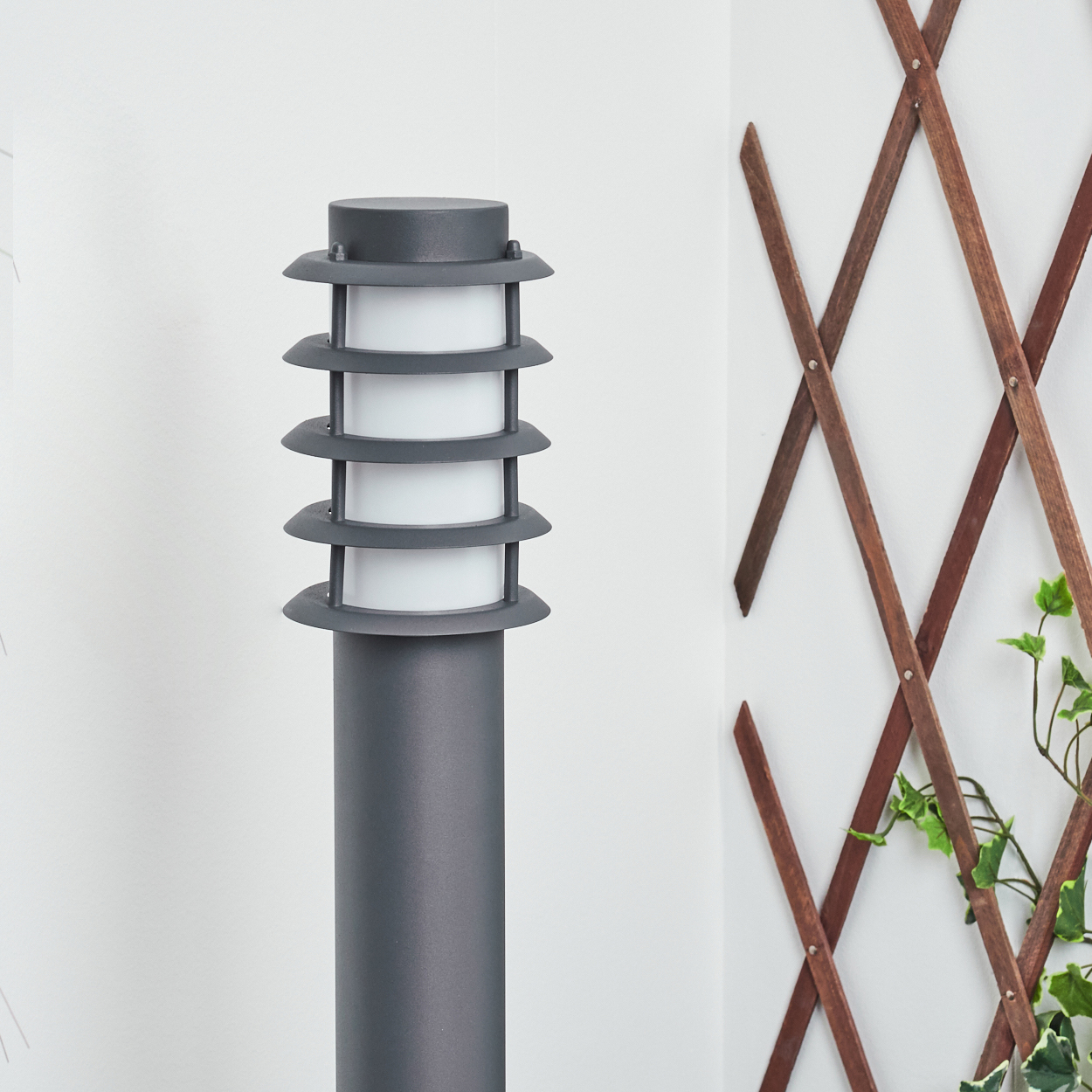 Graue, zylindrische Außenlampe mit gestapelten, horizontalen Lichtern. Holzgitter im Hintergrund.