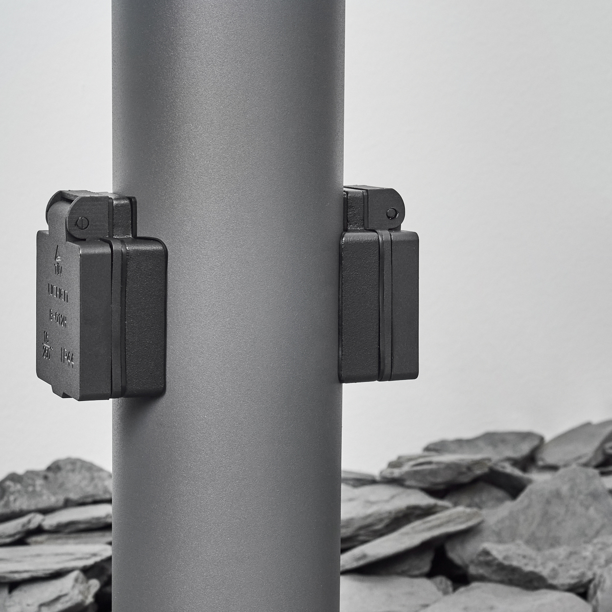 Nahaufnahme eines grauen Lampenpfostens mit zwei schwarzen Kästen. Steine im Hintergrund.