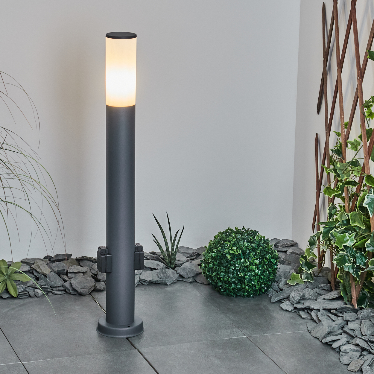 Ein moderner zylindrischer Lampenpfosten mit weißem Oberteil und grauem Körper, in einer Gartenszene.