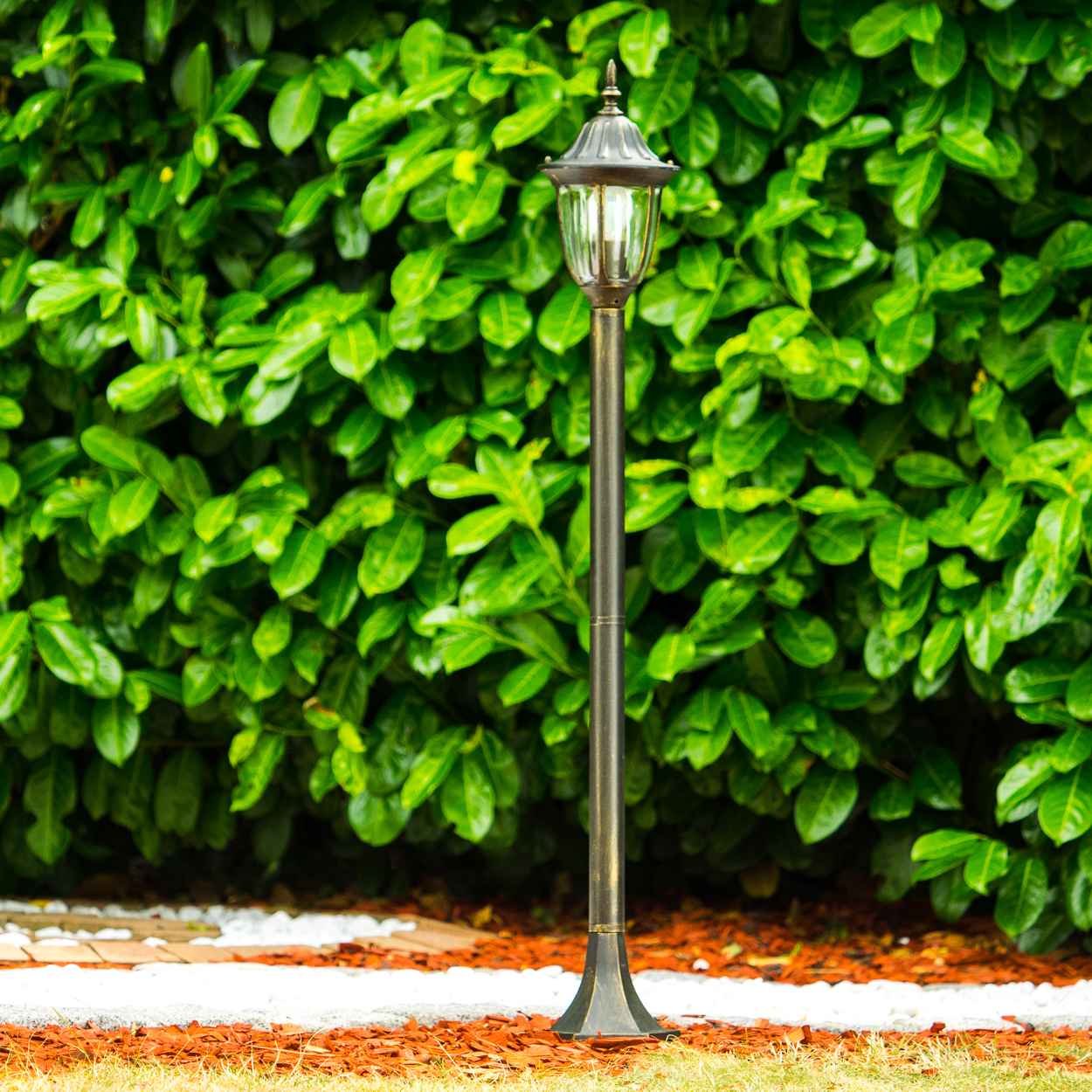 Eine bronzefarbene Außenlampe mit grünem Laub im Hintergrund, auf rotem Mulch und weißen Steinen.