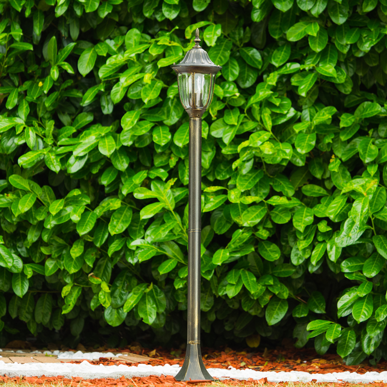 Eine Außenlampe vor einer grünen Hecke, in einem Garten mit weißen Steinen.
