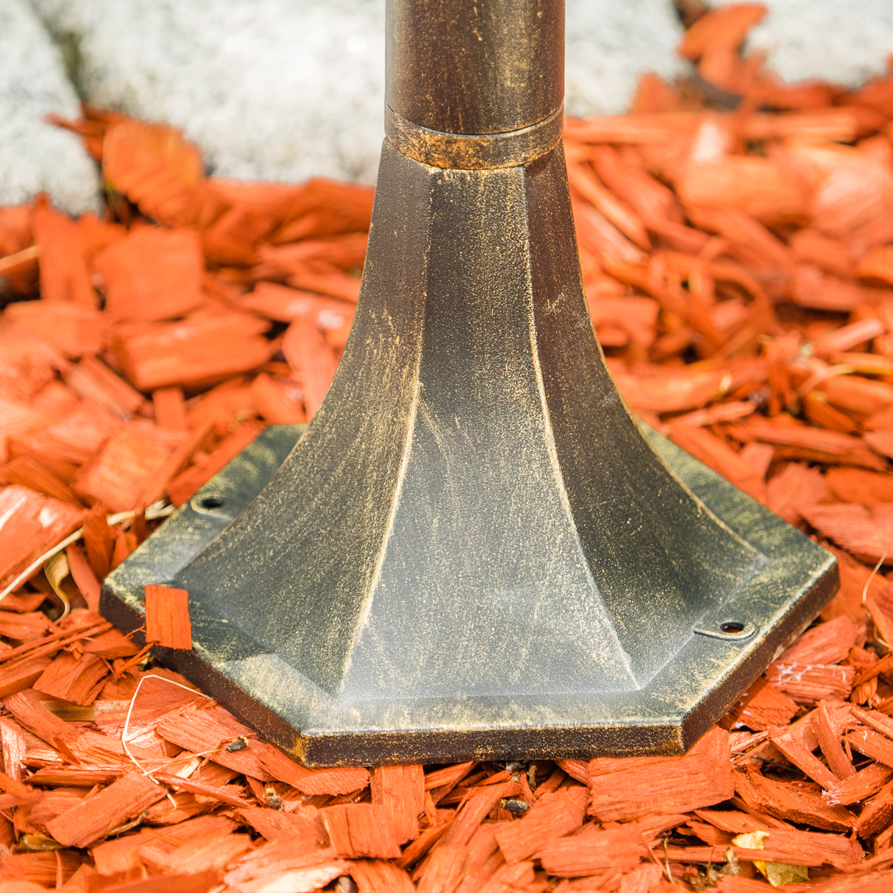 Nahaufnahme eines bronzefarbenen Lampenfußes in roter Mulch. Die Basis ist sechseckig.