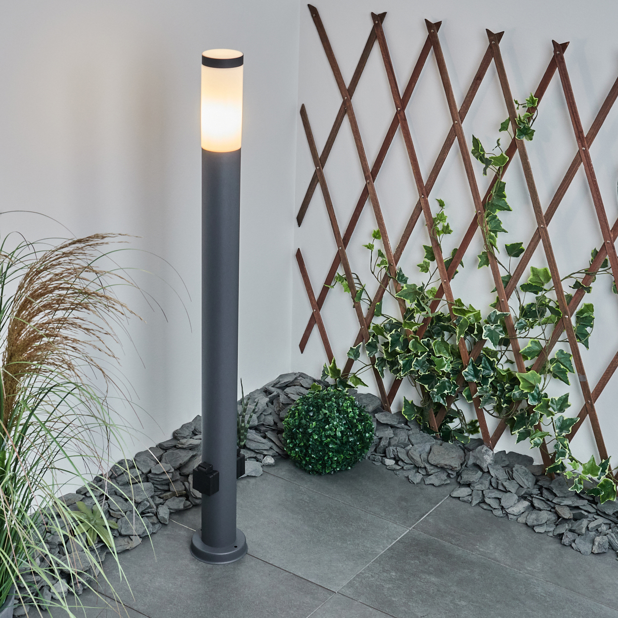 Eine graue Außenlampe mit leuchtender Spitze, neben Grün und einem Holzgitter.