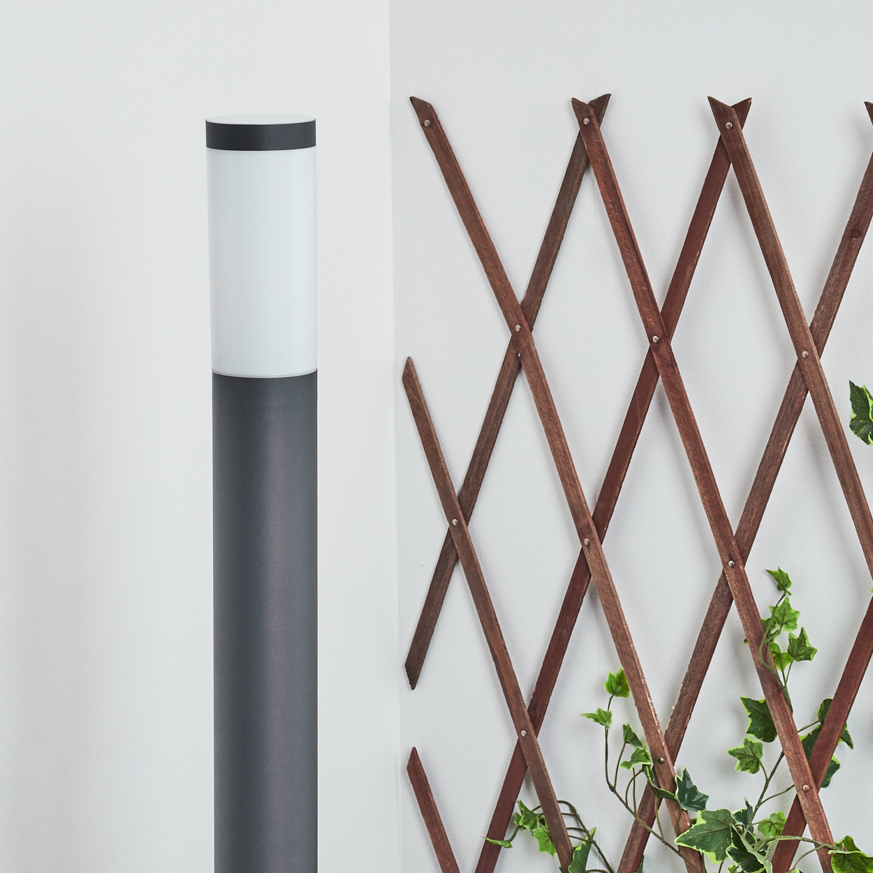 Ein grauer, zylindrischer Laternenpfahl neben einem hölzernen Spalier mit grünen Ranken.