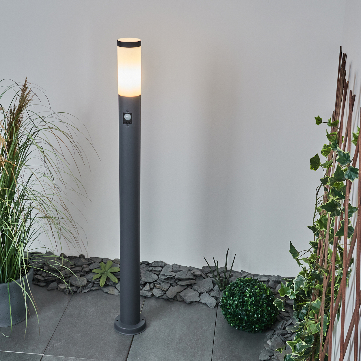 Graue Außenlampe mit Bewegungssensor. Die Lampe steht auf einem Steingrund neben Pflanzen und einem Holzgitter.