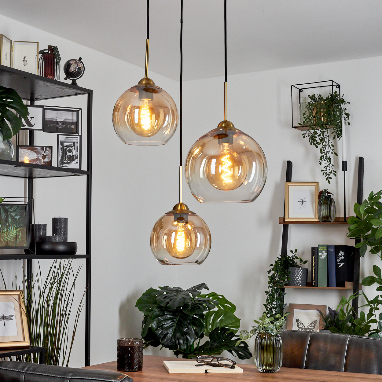 Drei Pendelleuchten über einem Holztisch, mit Pflanzen und Regalen im Hintergrund.