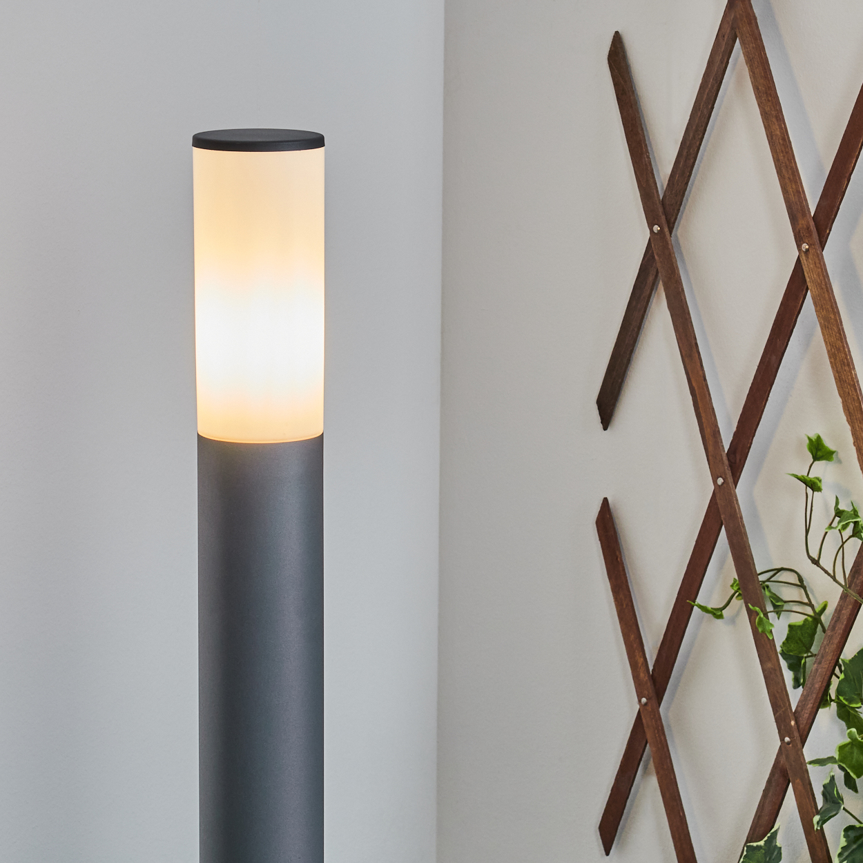 Beleuchteter grauer Lampenpfahl mit weißem Oberteil, neben einem Holzgitter vor weißer Wand.