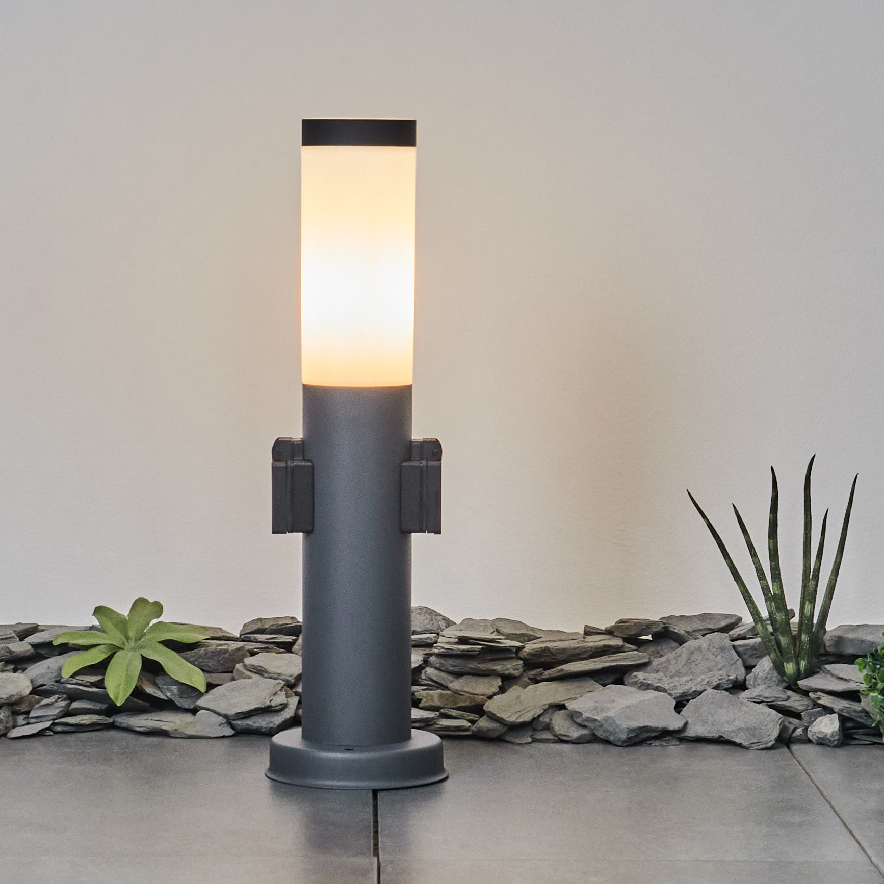Graue Laterne mit Licht an, umgeben von Steinen und Pflanzen. Weißer Wandhintergrund.