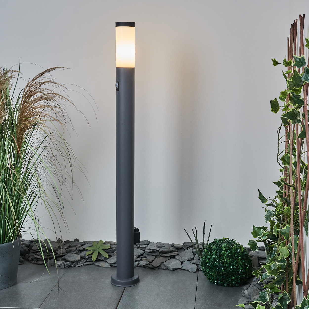 Graue Säulenlampe mit weißem Licht, Bewegungssensor und Steingrund. Grüne Pflanzen und weißer Wandhintergrund.