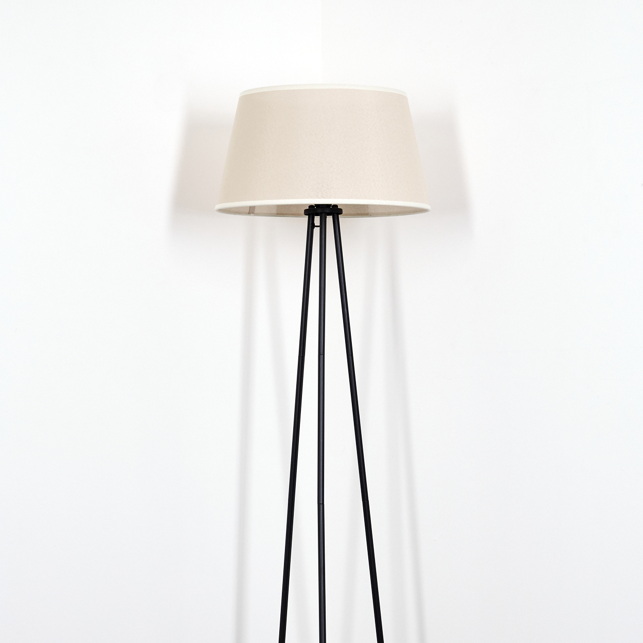 Stehlampe mit beigem Lampenschirm und schwarzen Dreibeinbeinen. Weißer Wandhintergrund.