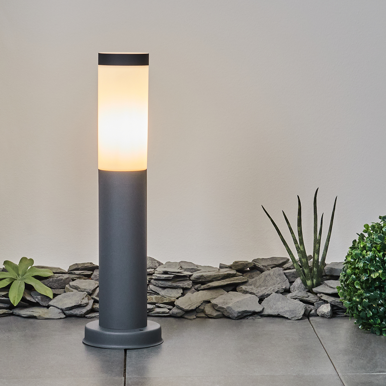 Beleuchtete graue Außenlampe mit weißem Oberteil auf einer grauen Terrasse. Grüne Pflanzen und Steine im Hintergrund.