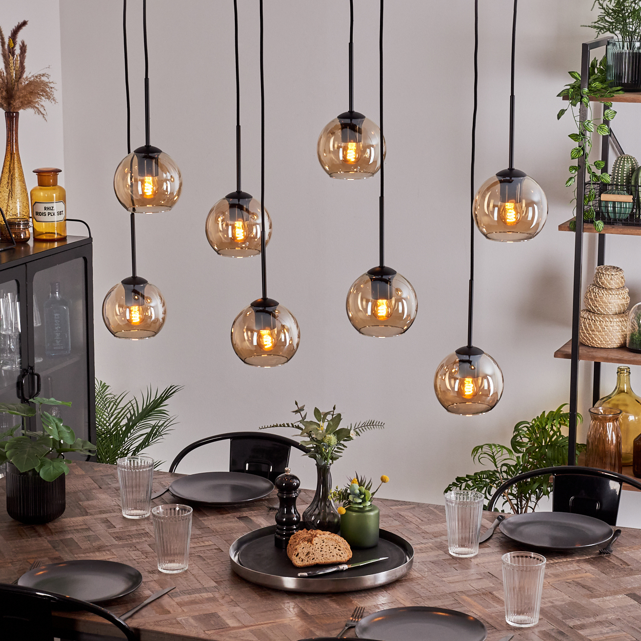 Acht bernsteinfarbene Glaskugelleuchten hängen über einem Holztisch, der für das Abendessen gedeckt ist, im Hintergrund Pflanzen.