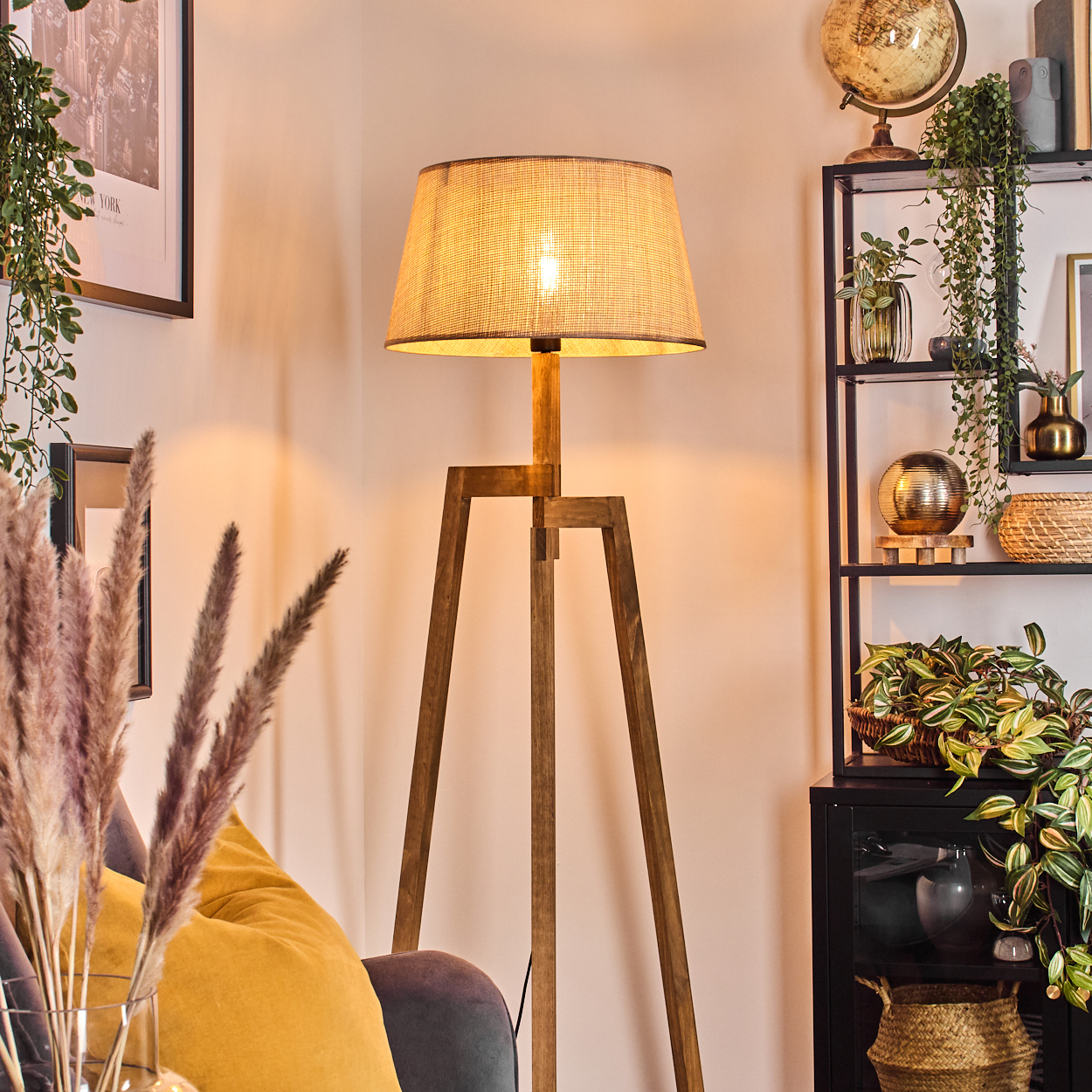 Beleuchtete Holz-Stativ-Stehleuchte mit beigem Schirm, in einem Wohnzimmer mit Möbeln und Pflanzen.