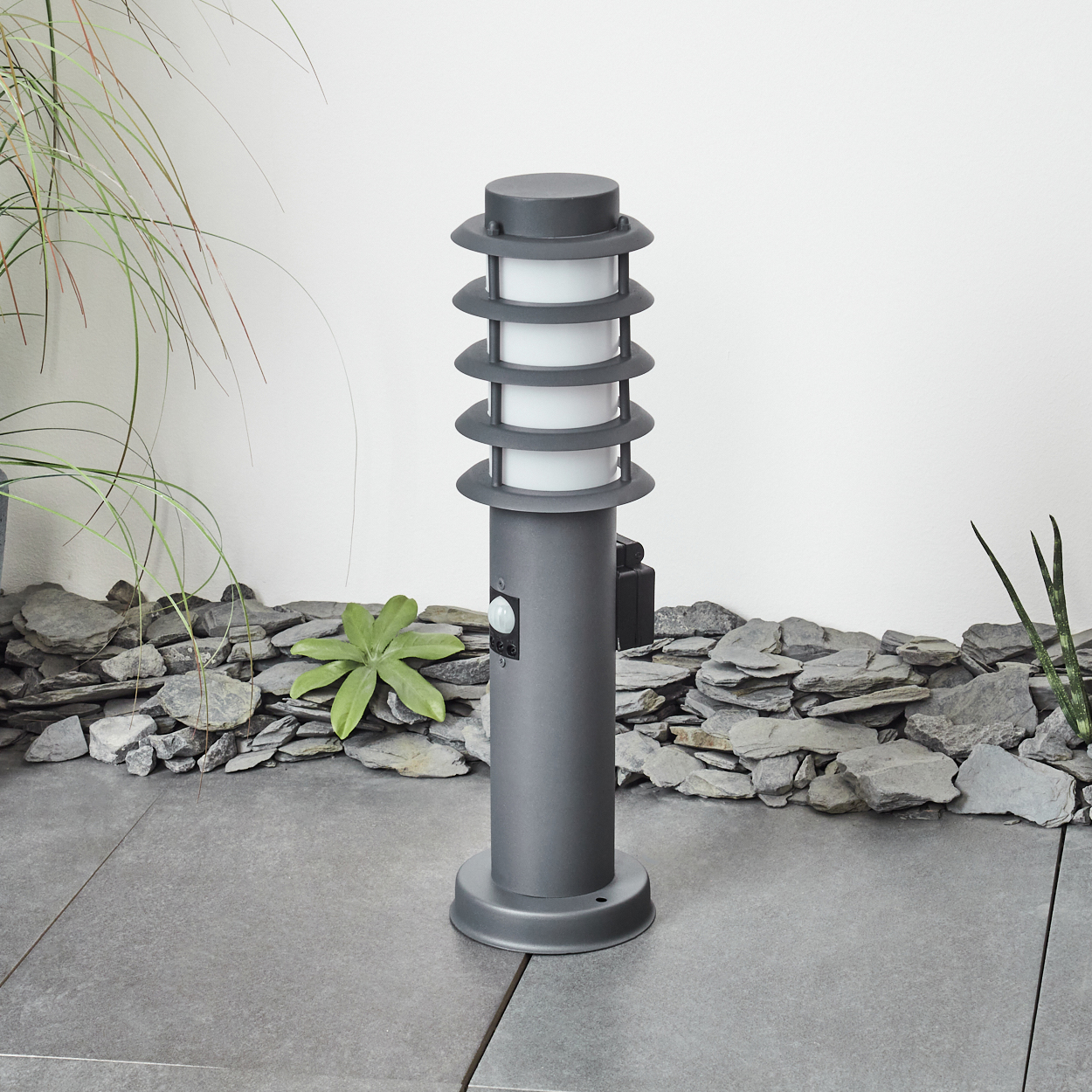 Graue Außenlampe mit gestapeltem Design, auf Pflastersteinen mit Felsen und Pflanzen.