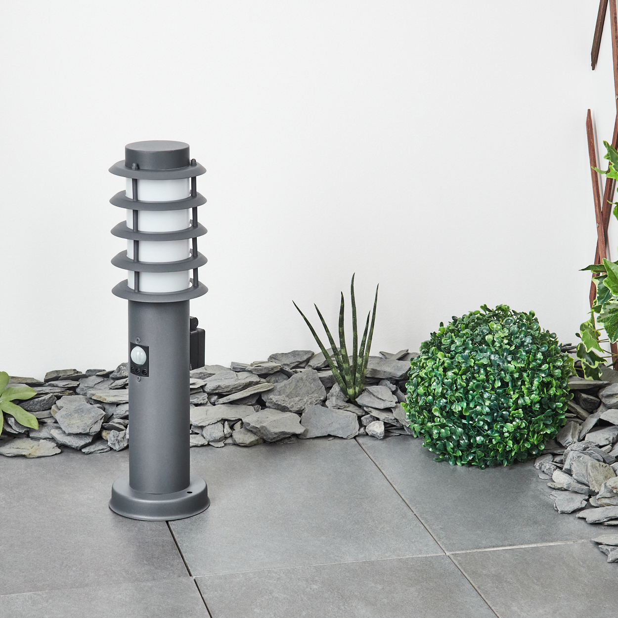 Graue Außenlampe mit gestapeltem Design, auf Pflastersteinen mit Felsen und Pflanzen.