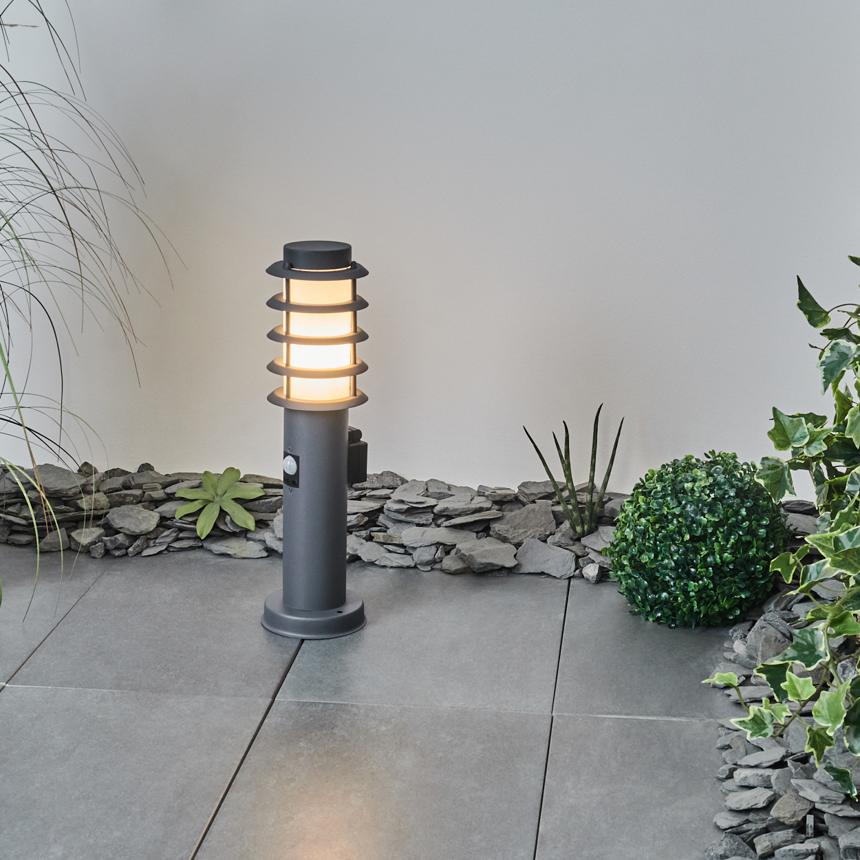 Eine graue Gartenlampe mit zylindrischem Körper. Die Lampe ist von Steinen und kleinen Pflanzen umgeben.