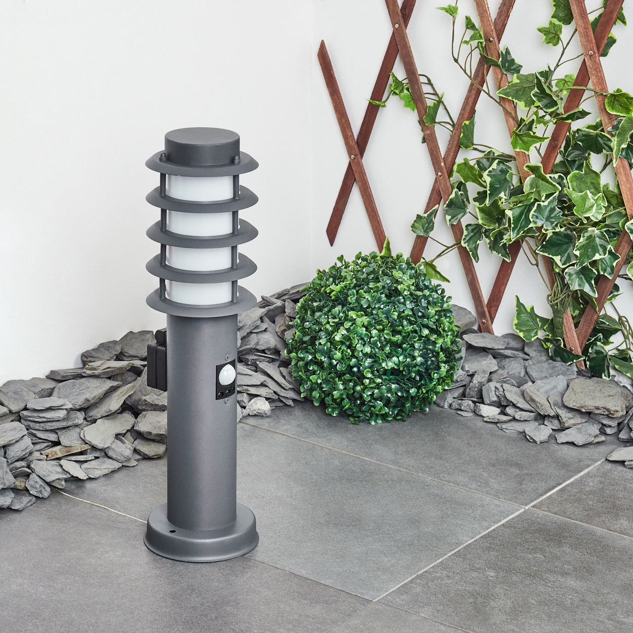 Grauer Außenmast mit Licht. Sensor und schwarze Steckdose sind sichtbar. Steinhintergrund.