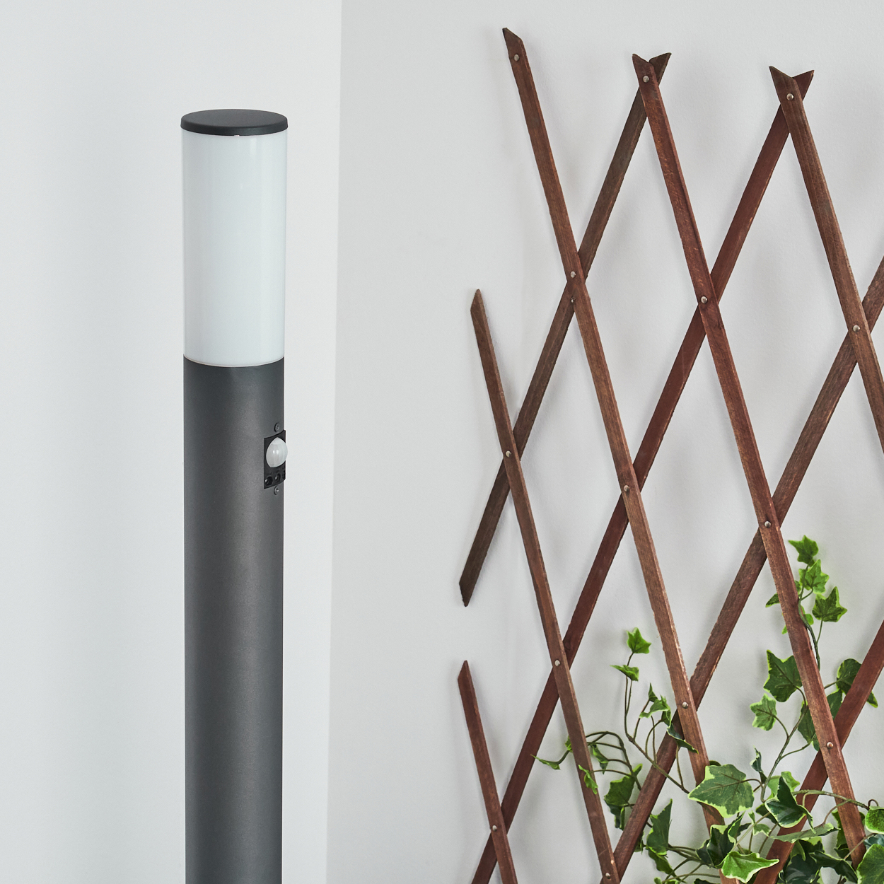 Graue Außenlampe und Holzspalier vor weißer Wand, mit Pflanzen.