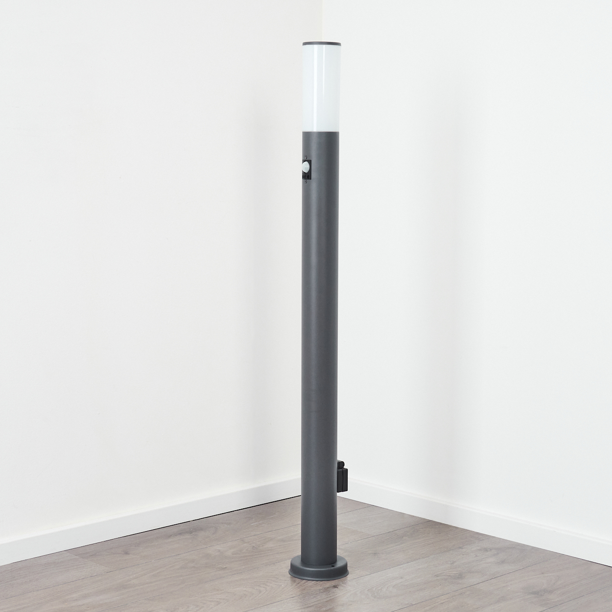 Dunkelgraue zylindrische Außenlampe mit weißem Oberteil, vor weißen Wänden und Holzboden.