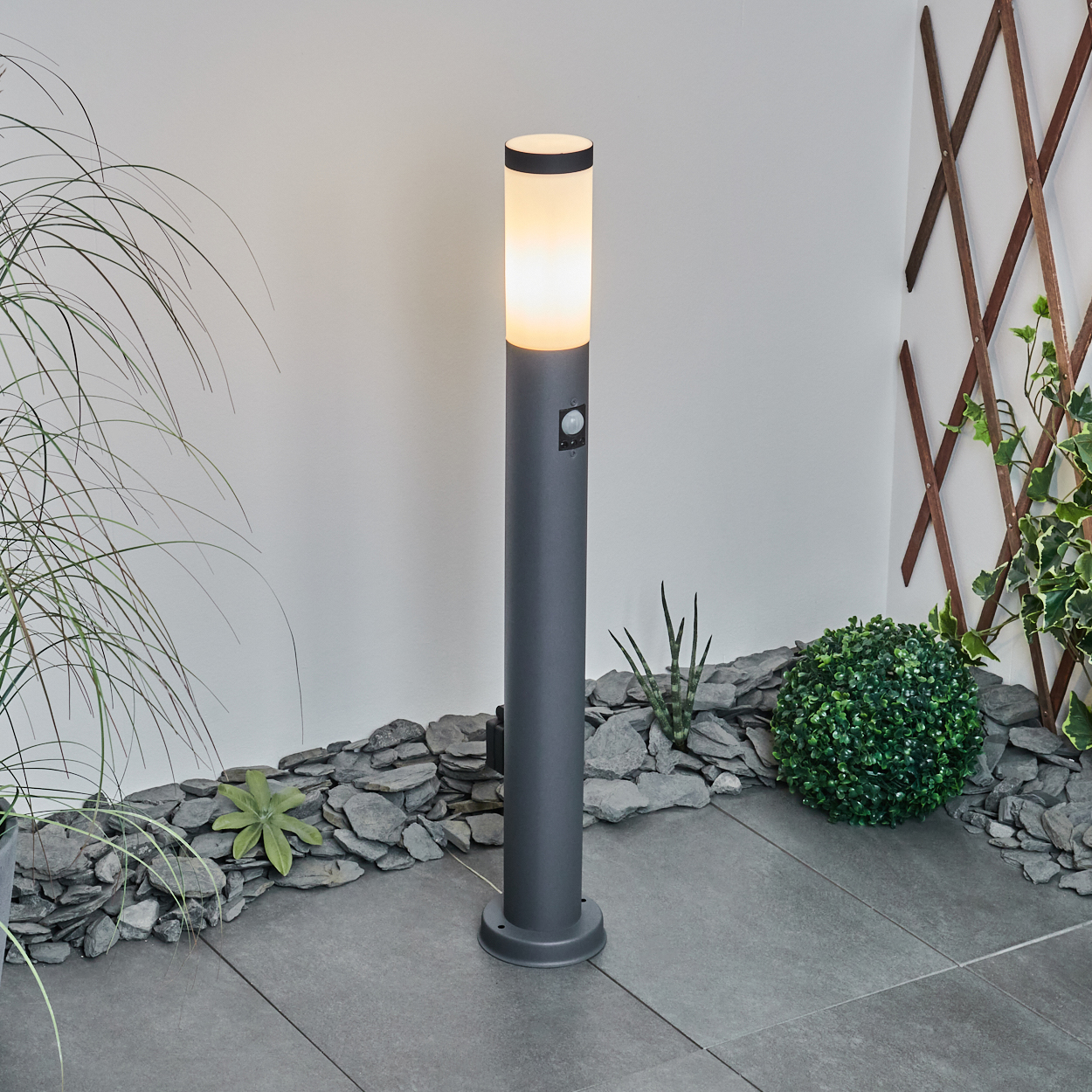 Leuchtende Außenlampe auf Steinfliesen. Hintergrund: Pflanzen, Steine und Holzgitter.