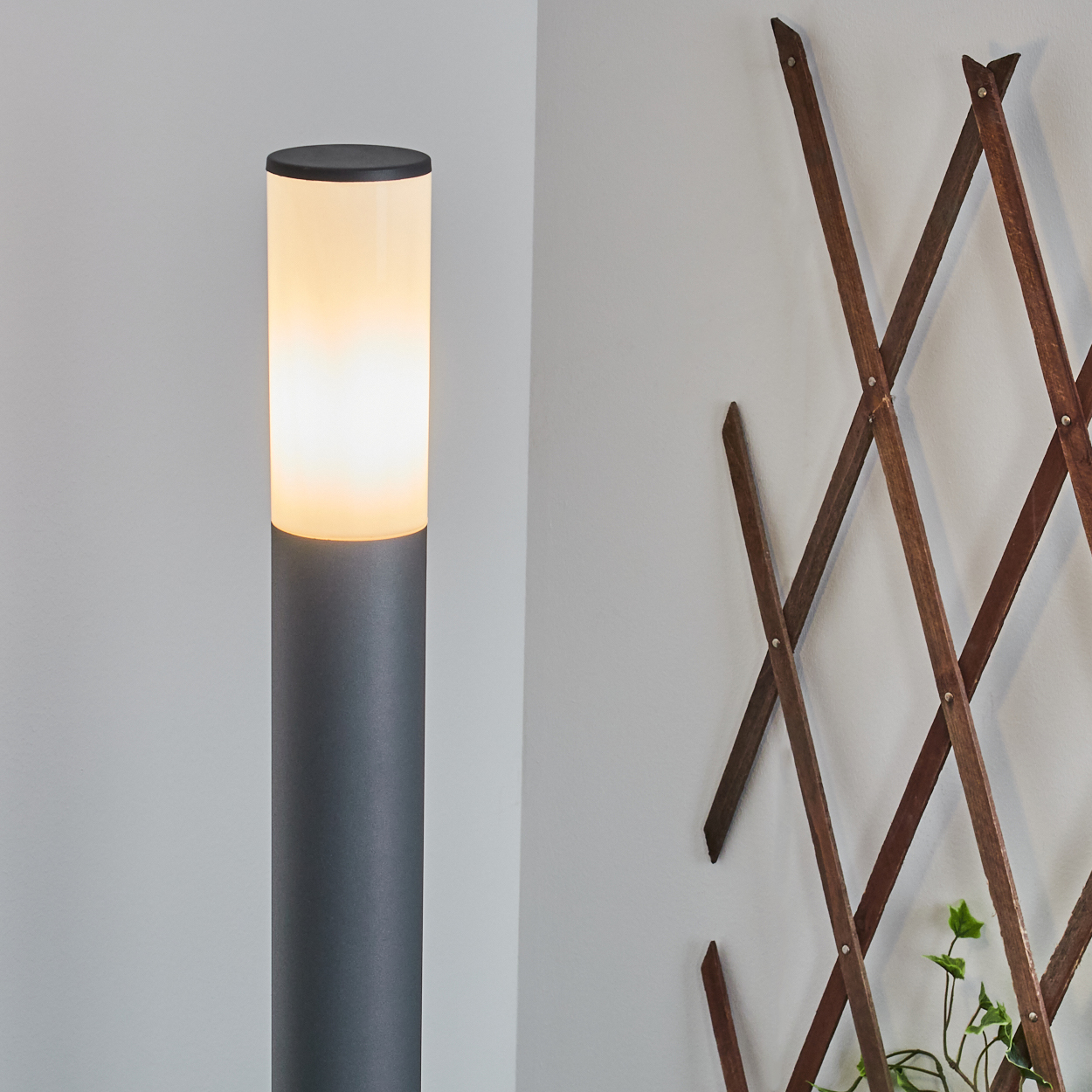 Hohe, beleuchtete graue Außenlampe vor einer weißen Wand mit einem Holzspalier und etwas Grün.