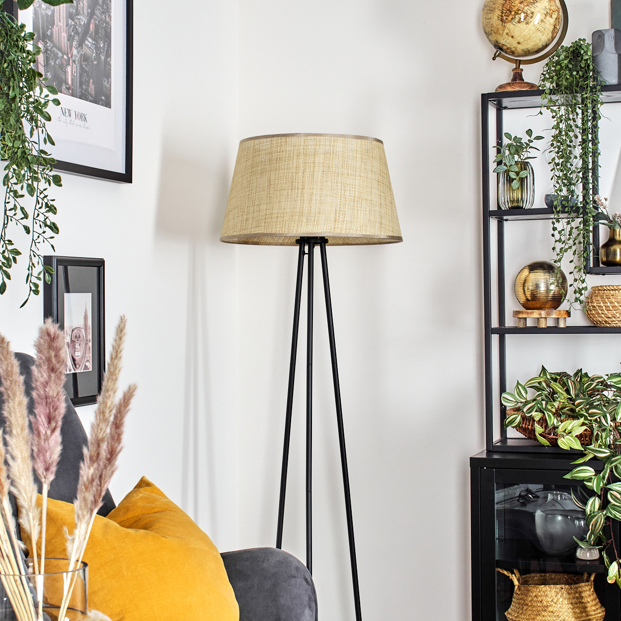 Stehlampe mit beigem Lampenschirm, schwarzem Rahmen, in einem Raum mit Sofa, Pflanzen und Globus.