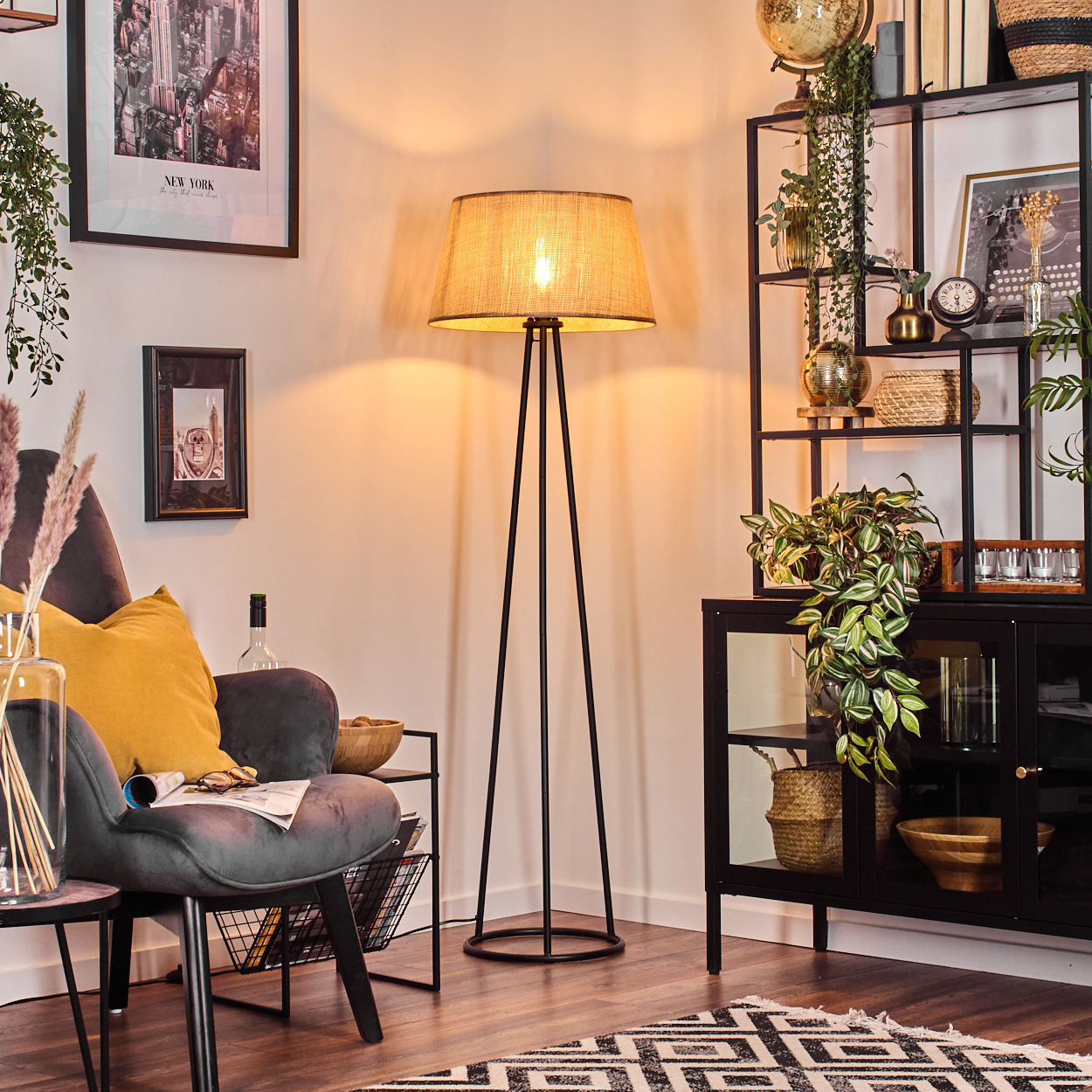 Stehlampe, beiger Lampenschirm, schwarzer Rahmen, in einem Raum mit Sofa, Tisch, Pflanzen und Kunstwerken. Es ist Tag.