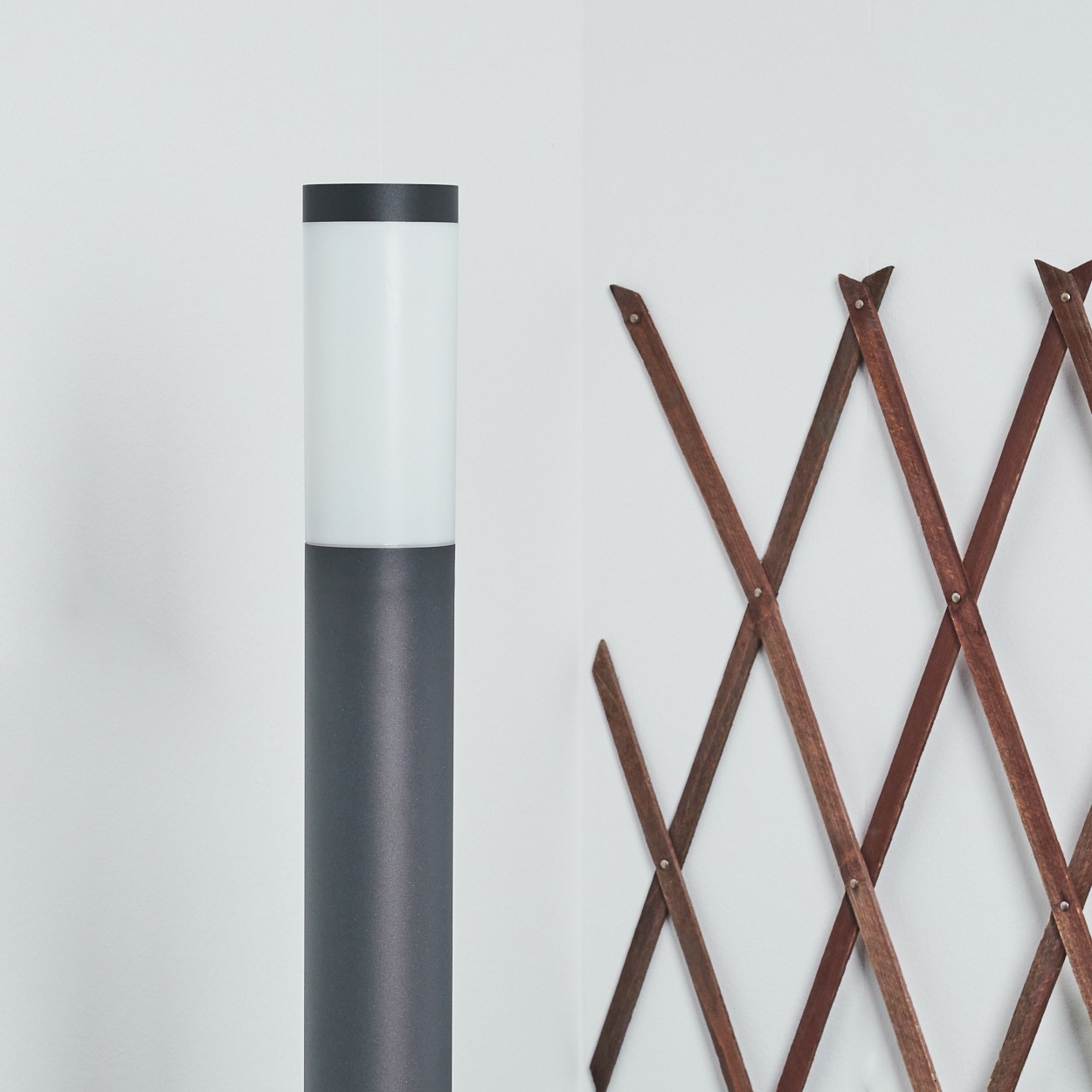 Eine Außenlampe mit grauem Pfahl und weißem, beleuchtetem Abschnitt, rechts ein Holzgitter.