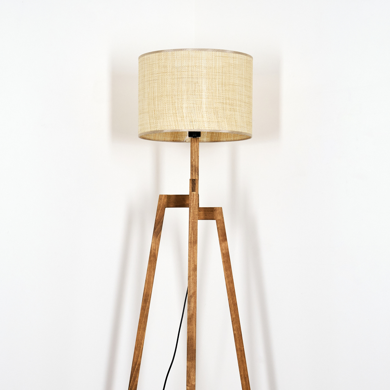 Holz-Stativ-Stehlampe mit beigem Lampenschirm. Steht vor einer weißen Wand.