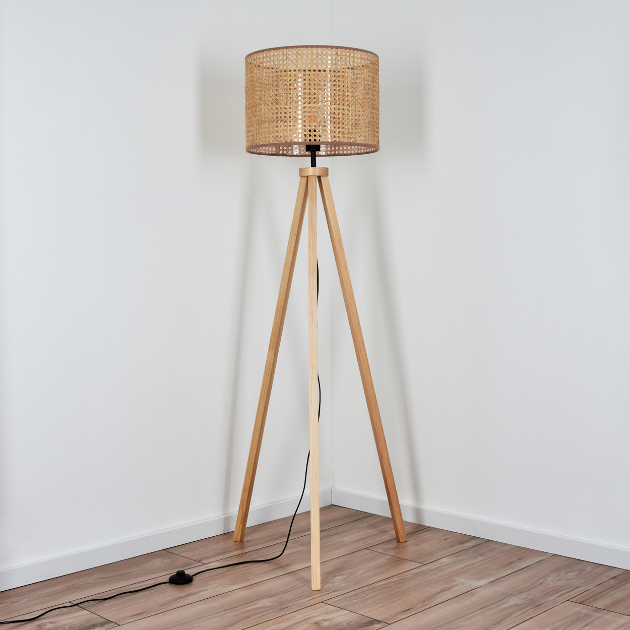 Dreibein-Stehlampe aus Holz mit gewebtem Lampenschirm. Helle Holzbeine. Weiße Wand, Holzboden. Eckaufstellung.