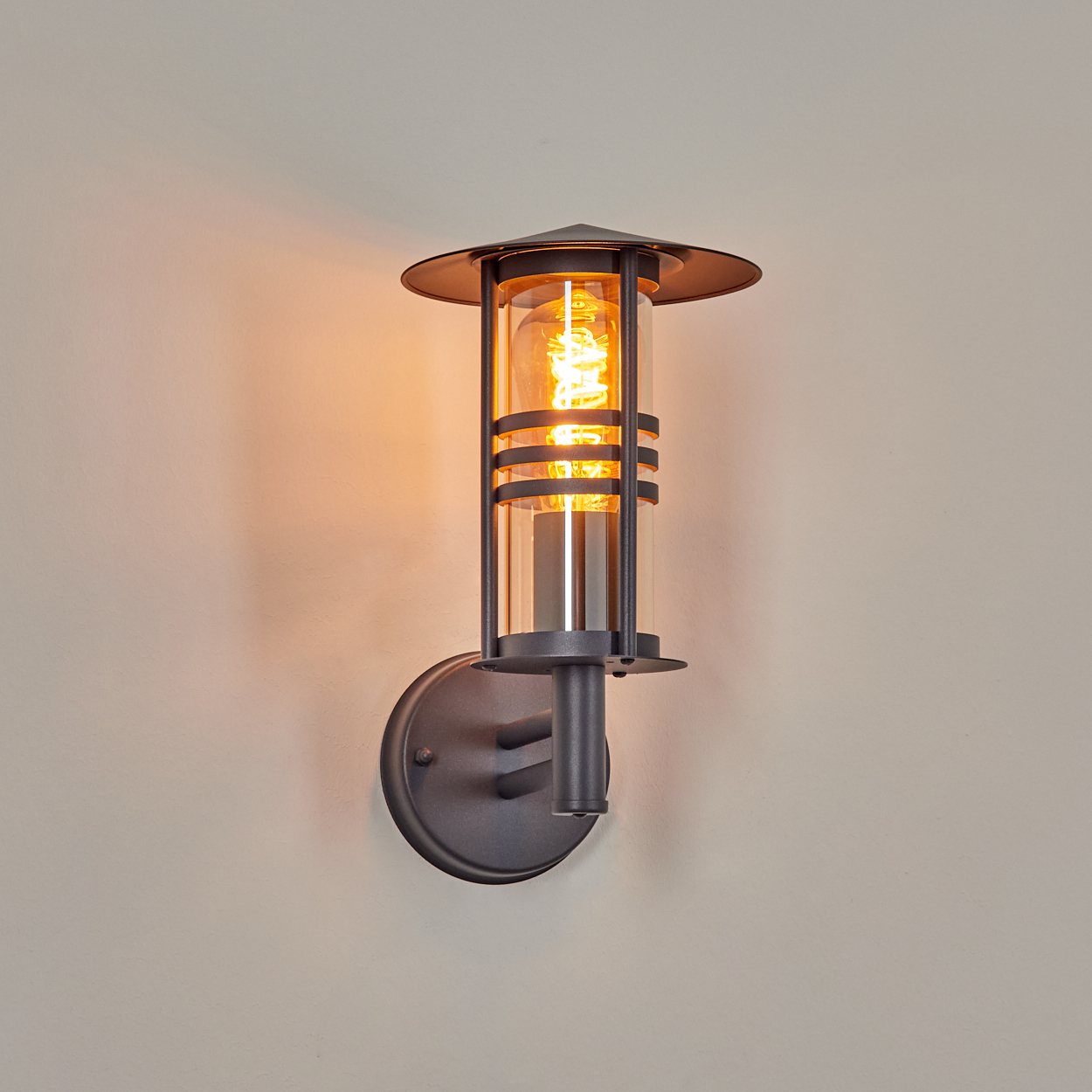 Wandleuchte, an einer Wand montiert. Schwarzer Metallrahmen, Glasröhre, bernsteinfarbene Glühbirne. Warmes Licht an beiger Wand.