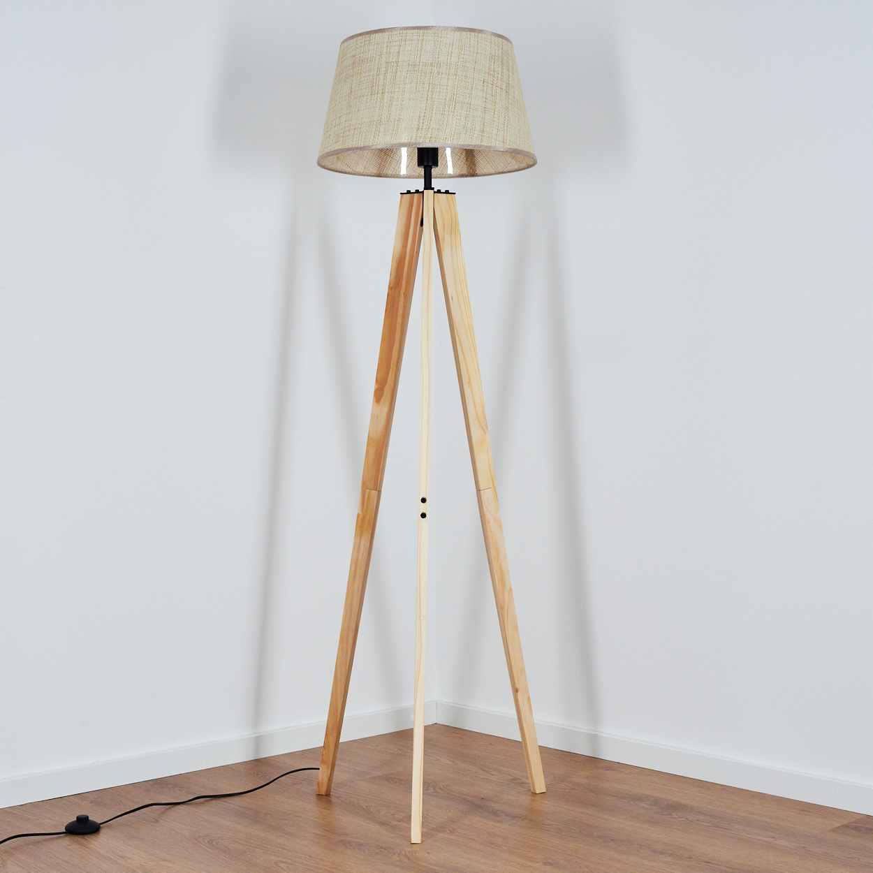 Eine hölzerne Stativ-Stehlampe in einer Zimmerecke. Die Lampe hat einen beigefarbenen Lampenschirm.