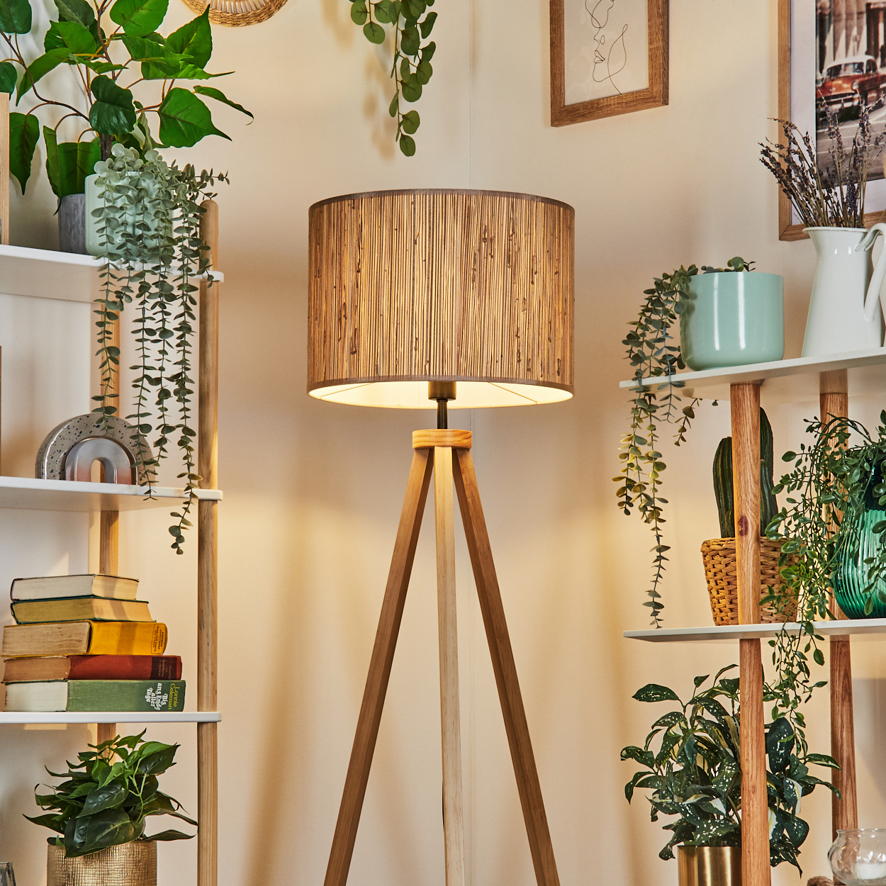Wohnzimmer mit einer hölzernen Stativlampe, Pflanzen und Regalen. Die Lampe hat einen gewebten Schirm.