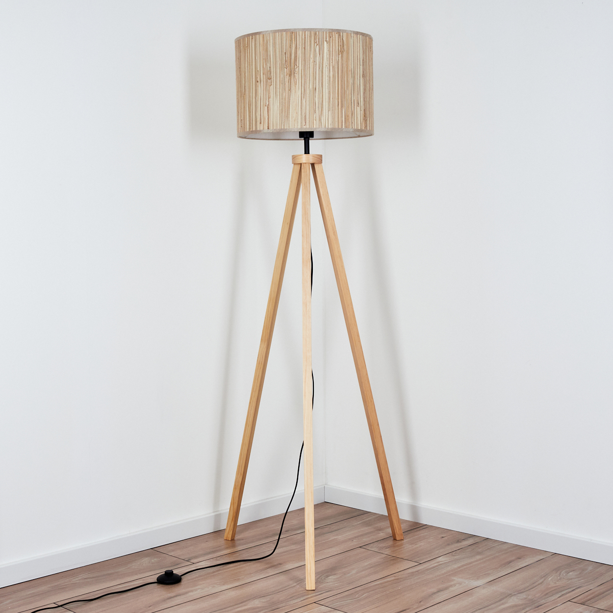 Stehlampe mit hölzernen Stativbeinen und gewebtem Lampenschirm. In einer Ecke neben Holzboden.