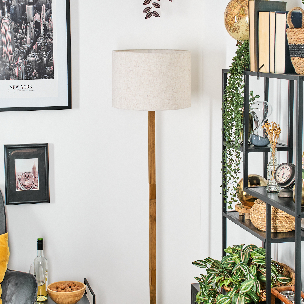 Holz-Stehlampe mit beigem Schirm. Im Hintergrund schwarzes Regal, Kunst und Pflanzen.