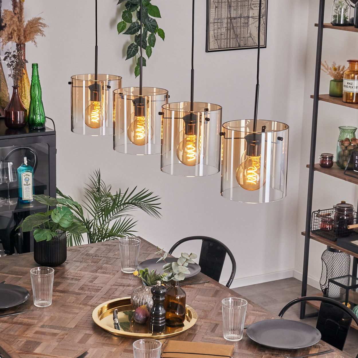 Vier Pendelleuchten mit bernsteinfarbenen Glasschirmen hängen über einem Holztisch. Pflanzen und Dekoration im Hintergrund.