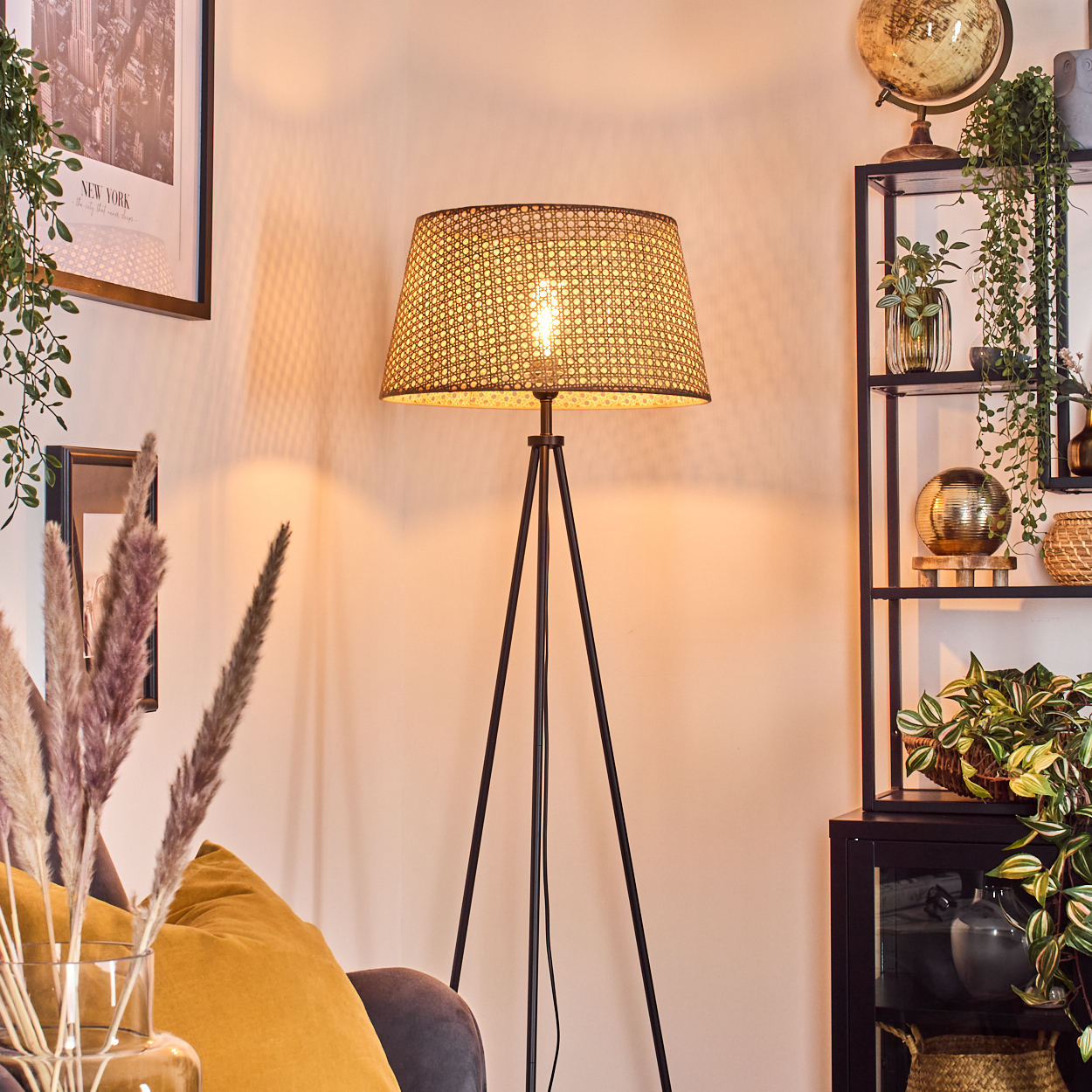 Stehlampe mit gewebtem Schirm in einem Raum. Sofa, Pflanzen und Deko im Hintergrund.
