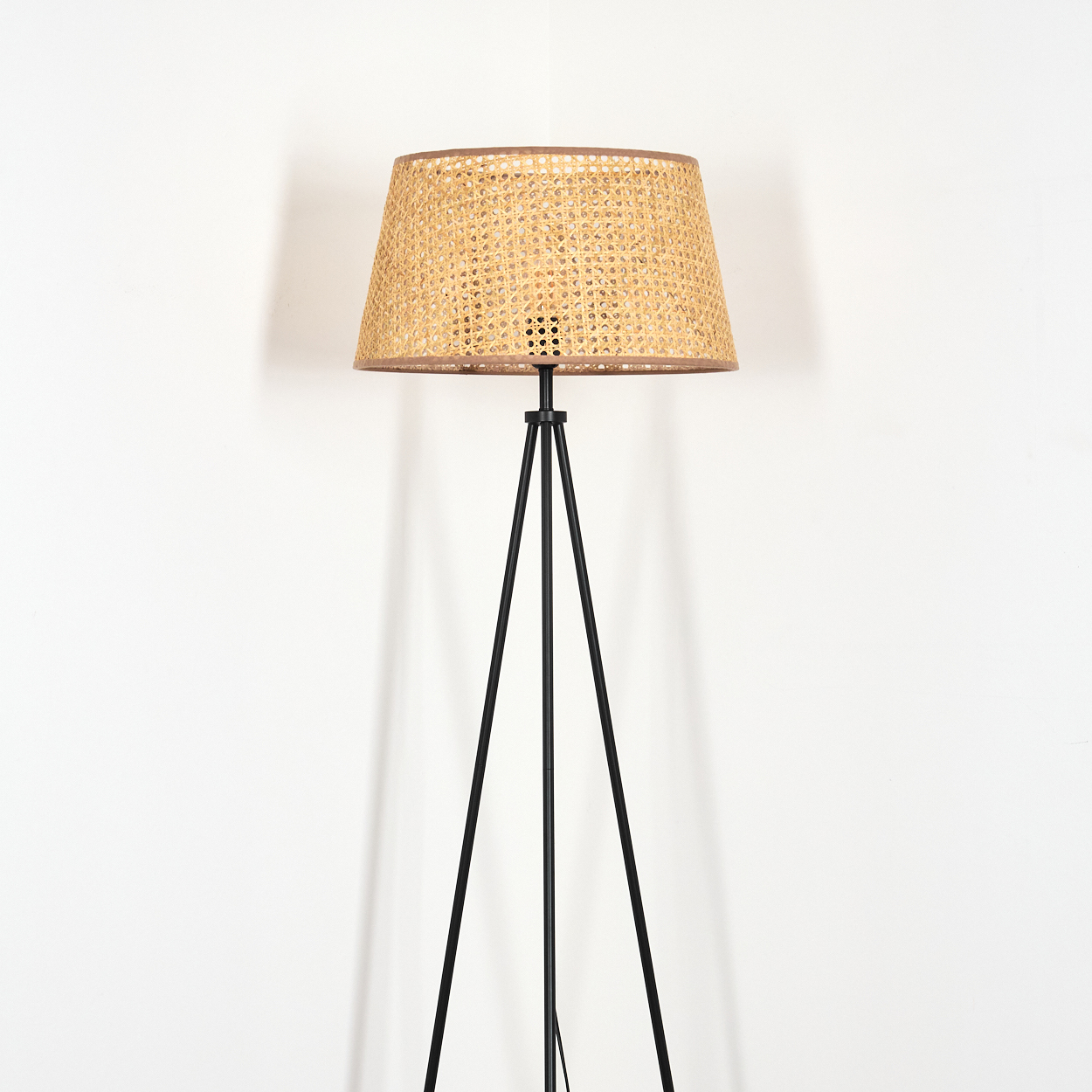 Eine Stehlampe mit beigem Rattan-Lampenschirm und schwarzem Dreibein-Sockel, vor weißem Hintergrund.