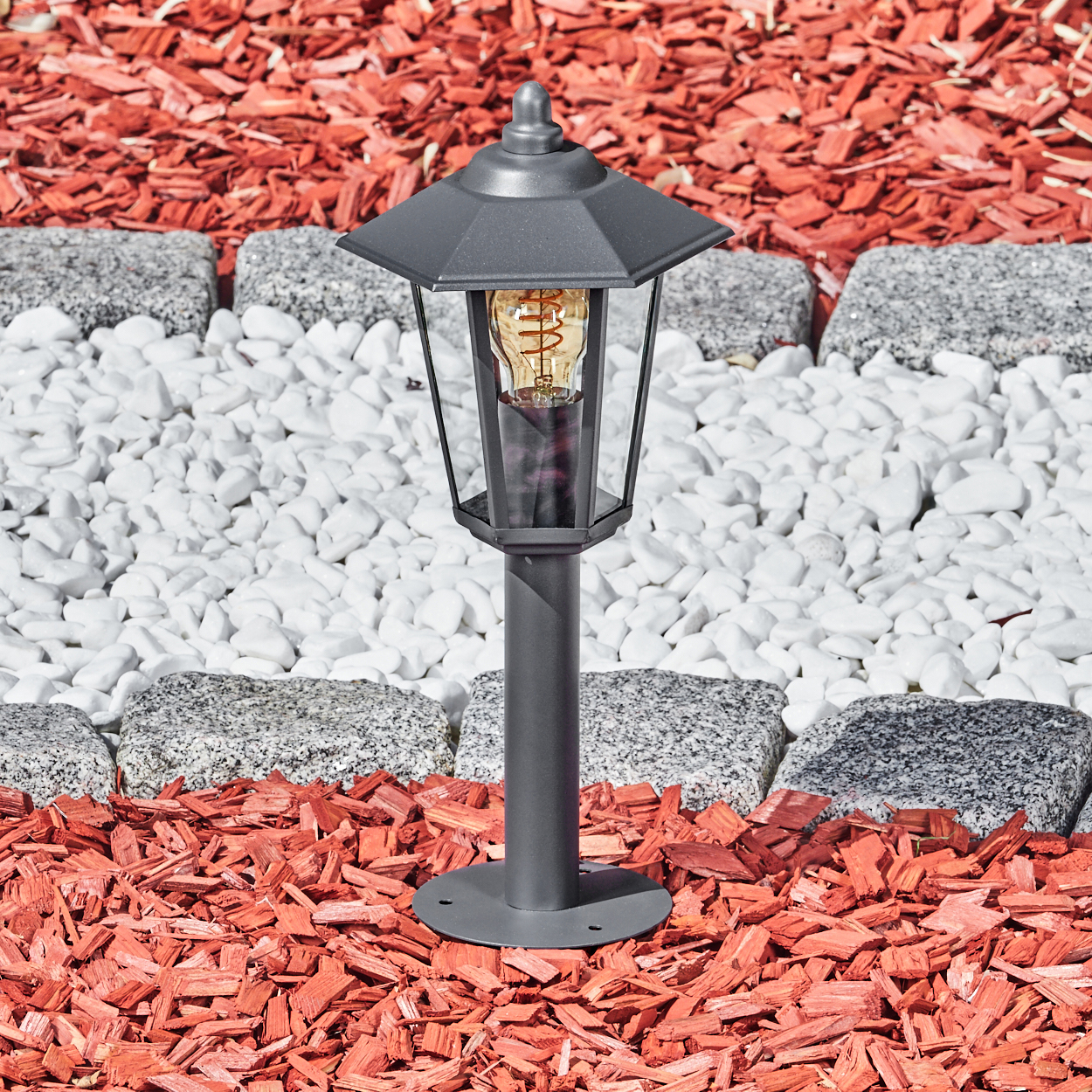 Graue Außenlaterne mit Glühbirne. Sie steht auf roter Mulch mit einem weiß-grauen Steinweg.