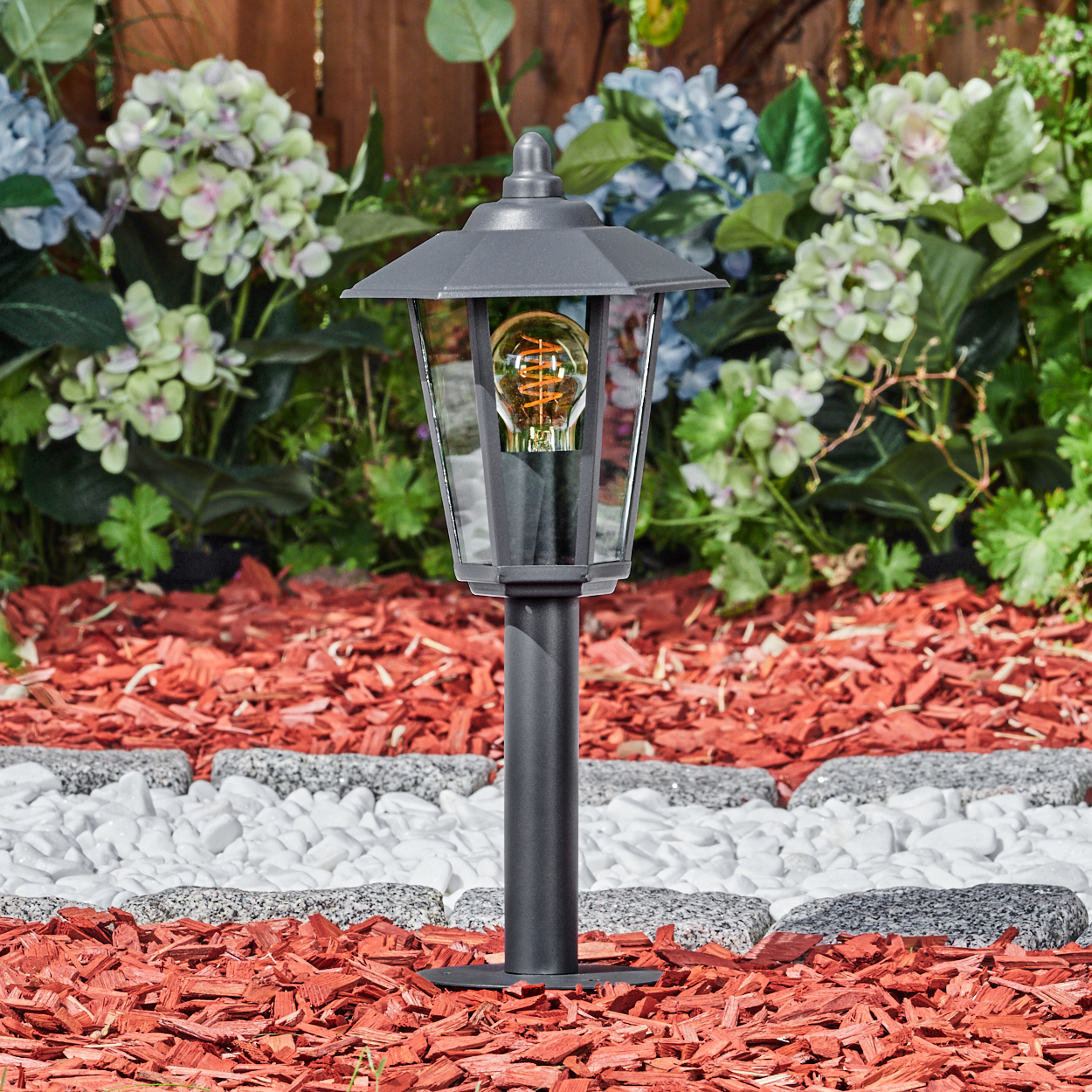 Outdoor-Laternenpfahl mit dunkelgrauer Oberfläche, Glaseinfassung und dekorativem Aufsatz, in einem Garten.