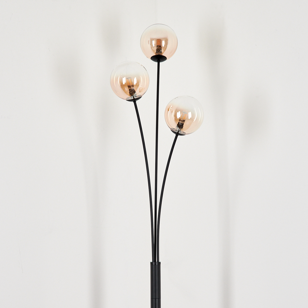 Schwarze Stehlampe mit drei bernsteinfarbenen Glaskugelschirmen, schlichtes Design vor weißer Wand.